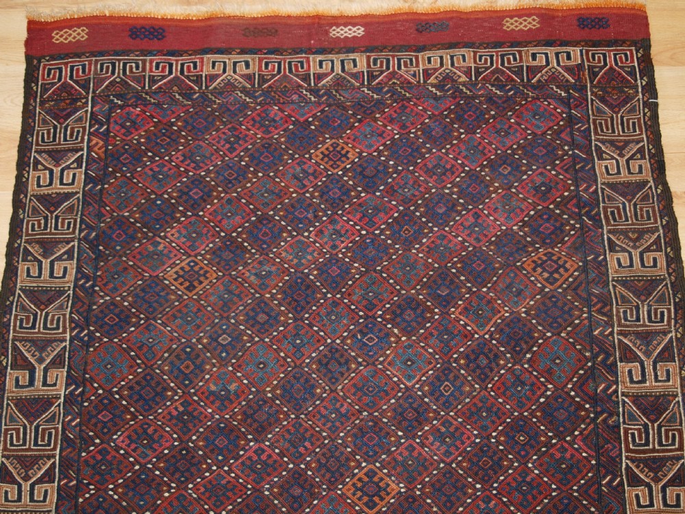 A long handwoven rectangular Kurdish Soumak kilim rug featuring a dense red and blue diamond lattice pattern framed by a geometric meander border. View IV