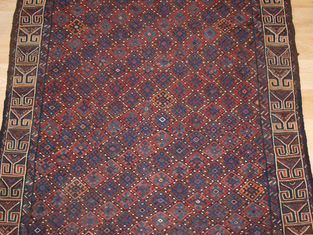 A long handwoven rectangular Kurdish Soumak kilim rug featuring a dense red and blue diamond lattice pattern framed by a geometric meander border. View VI