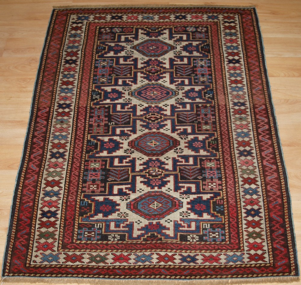 An antique Caucasian hand-knotted wool rug with geometric Lesghi star medallions on a cream field surrounded by patterned borders in red, navy, and ivory.