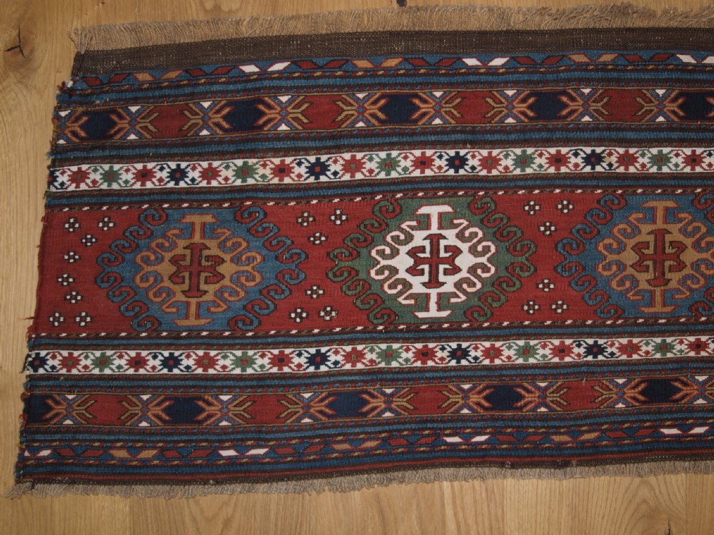 A rectangular hand-woven antique Caucasian Soumak wool panel featuring horizontal bands with geometric diamond medallions in terracotta red, navy blue, ivory, and green. View II