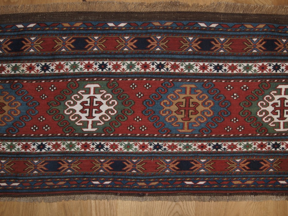 A rectangular hand-woven antique Caucasian Soumak wool panel featuring horizontal bands with geometric diamond medallions in terracotta red, navy blue, ivory, and green. View III