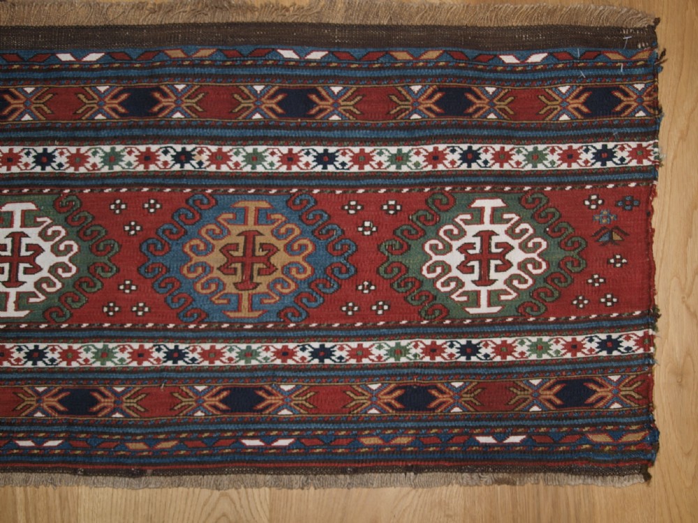 A rectangular hand-woven antique Caucasian Soumak wool panel featuring horizontal bands with geometric diamond medallions in terracotta red, navy blue, ivory, and green. View IV