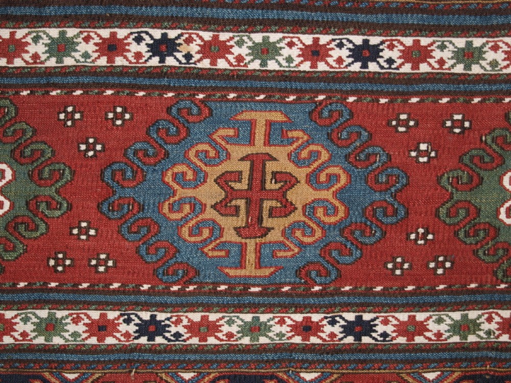 A rectangular hand-woven antique Caucasian Soumak wool panel featuring horizontal bands with geometric diamond medallions in terracotta red, navy blue, ivory, and green. View V