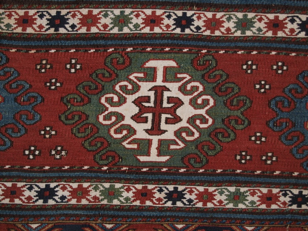 A rectangular hand-woven antique Caucasian Soumak wool panel featuring horizontal bands with geometric diamond medallions in terracotta red, navy blue, ivory, and green. View VI