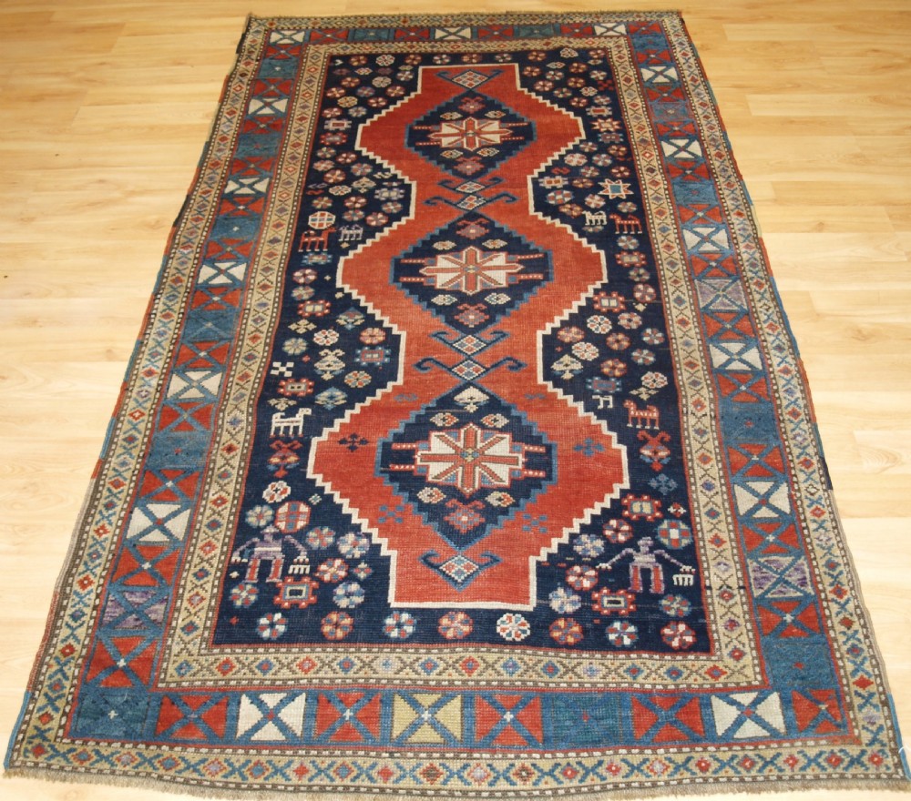 An antique hand-knotted Caucasian wool rug featuring red geometric diamond medallions on an indigo blue field with small human and animal figures.