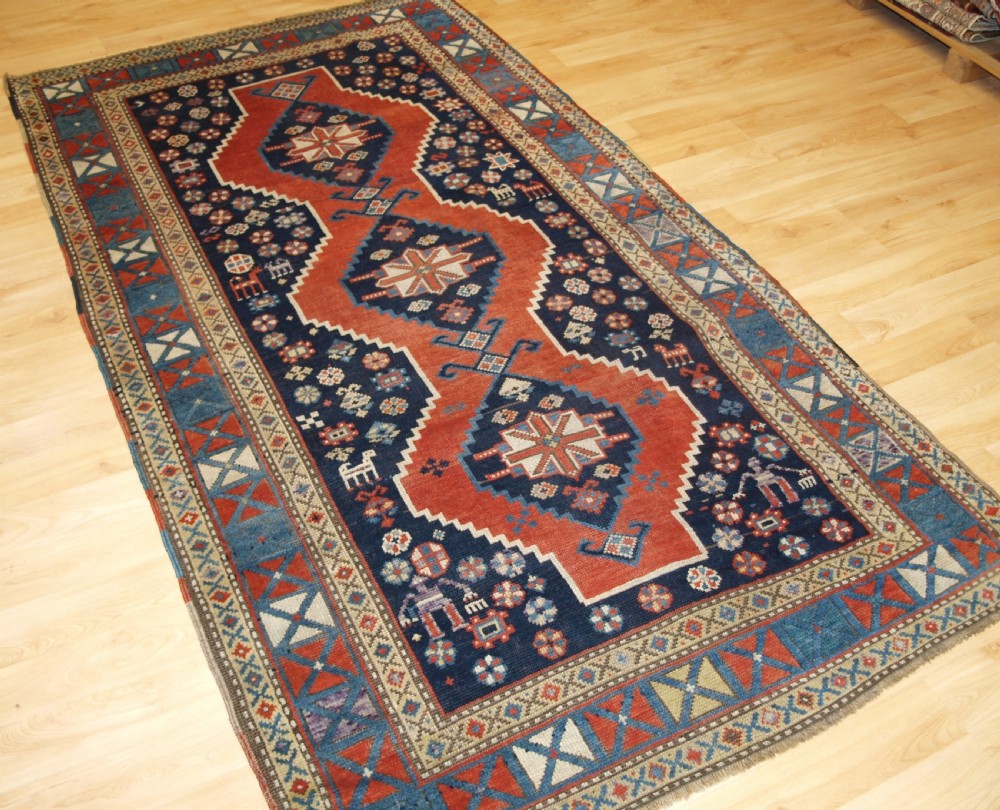 An antique hand-knotted Caucasian wool rug featuring red geometric diamond medallions on an indigo blue field with small human and animal figures. View II