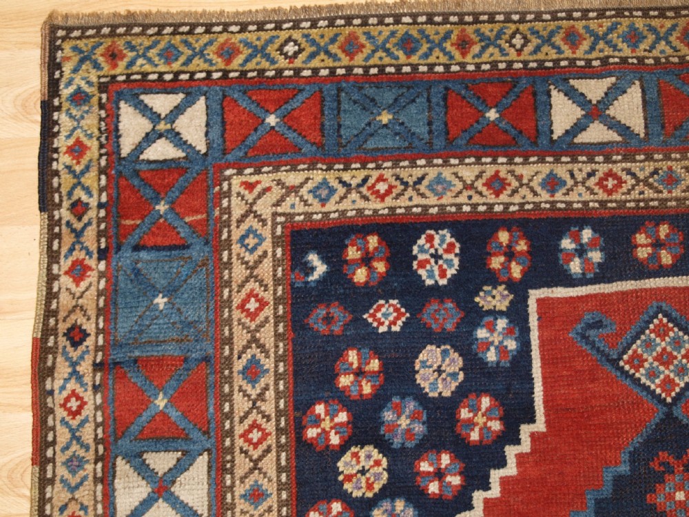 An antique hand-knotted Caucasian wool rug featuring red geometric diamond medallions on an indigo blue field with small human and animal figures. View III