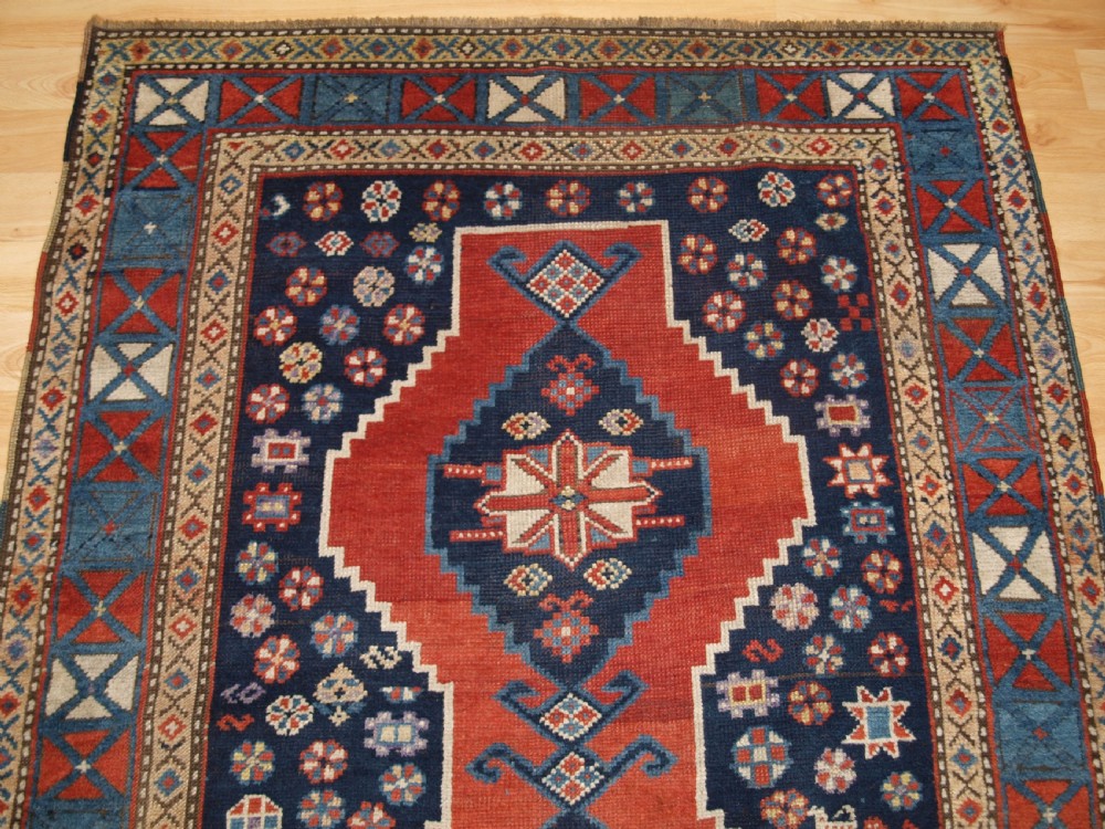 An antique hand-knotted Caucasian wool rug featuring red geometric diamond medallions on an indigo blue field with small human and animal figures. View IV
