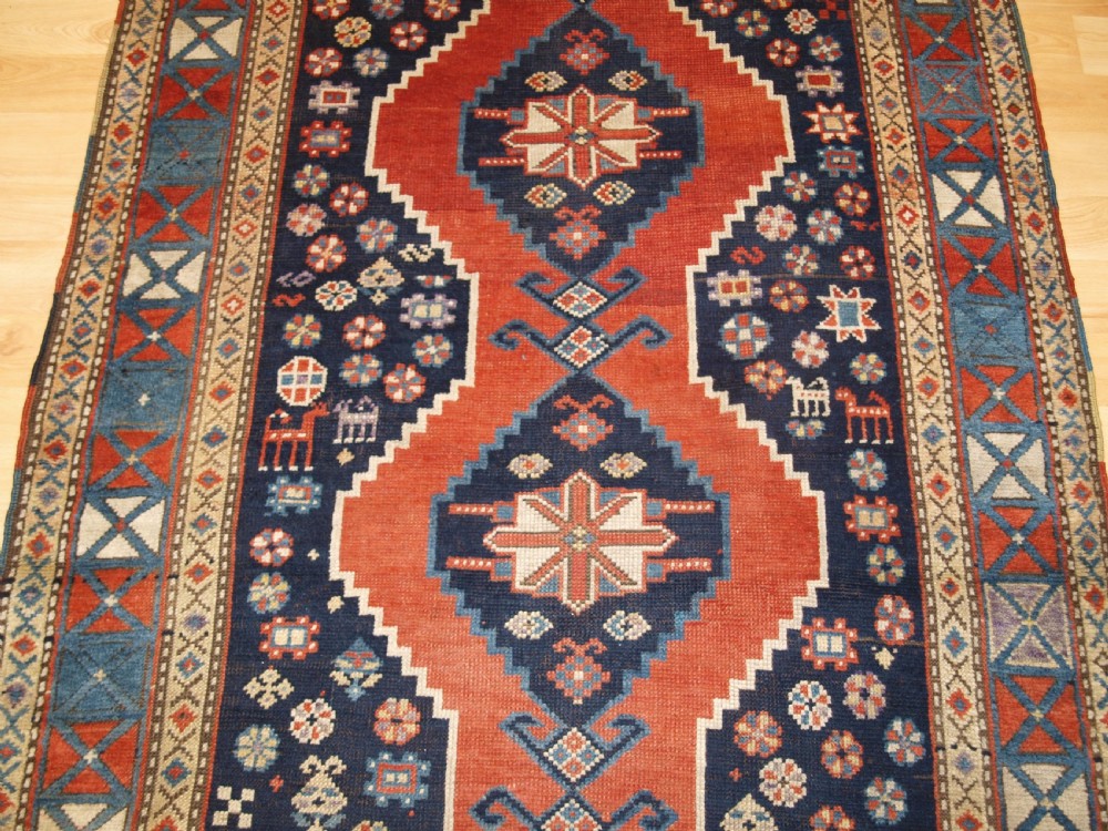An antique hand-knotted Caucasian wool rug featuring red geometric diamond medallions on an indigo blue field with small human and animal figures. View V