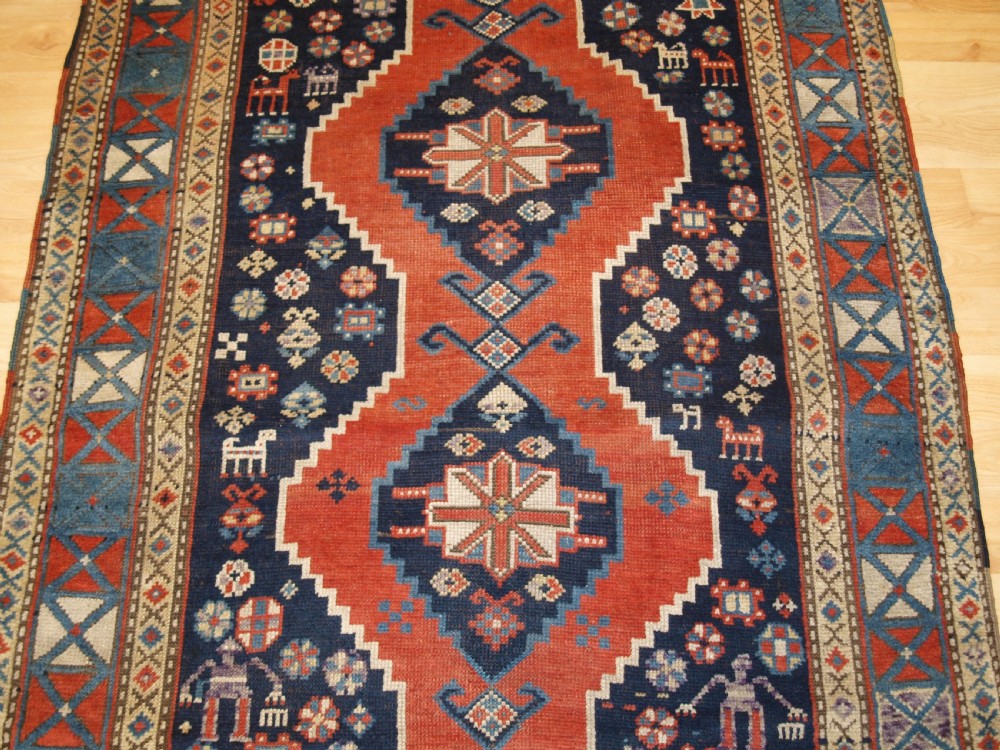 An antique hand-knotted Caucasian wool rug featuring red geometric diamond medallions on an indigo blue field with small human and animal figures. View VI