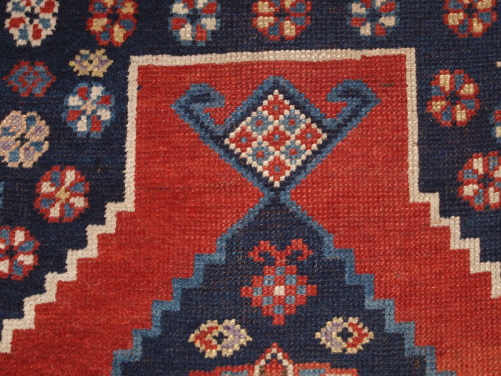 An antique hand-knotted Caucasian wool rug featuring red geometric diamond medallions on an indigo blue field with small human and animal figures. View VII