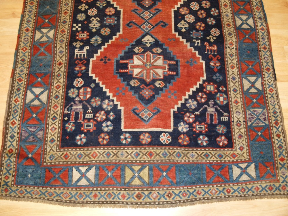 An antique hand-knotted Caucasian wool rug featuring red geometric diamond medallions on an indigo blue field with small human and animal figures. View VIII