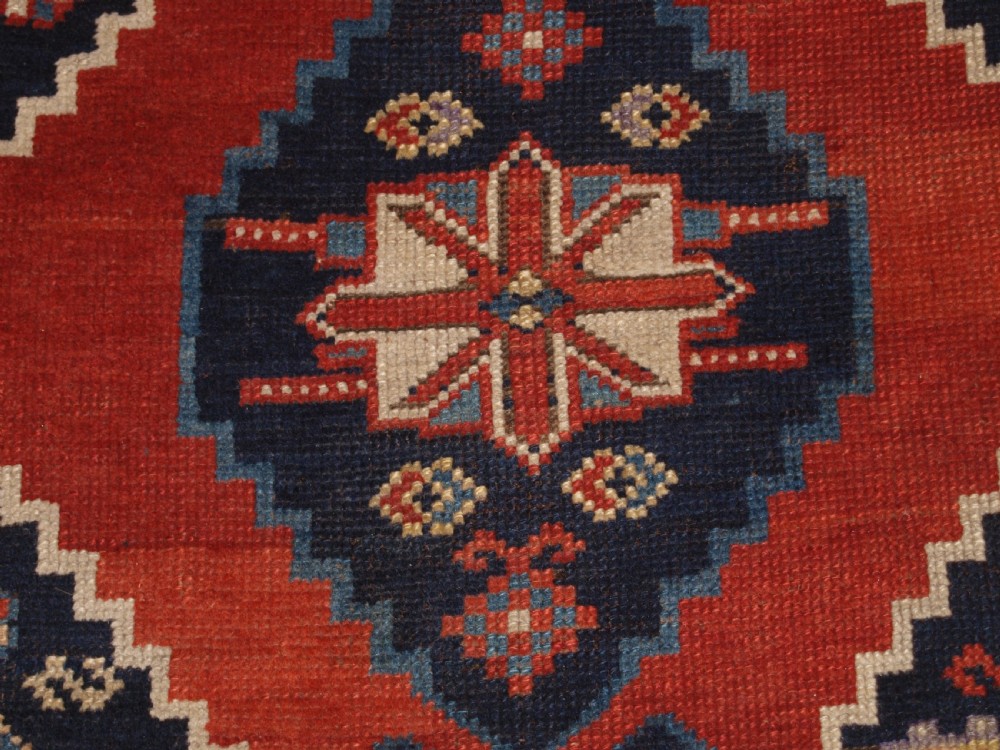 An antique hand-knotted Caucasian wool rug featuring red geometric diamond medallions on an indigo blue field with small human and animal figures. View IX