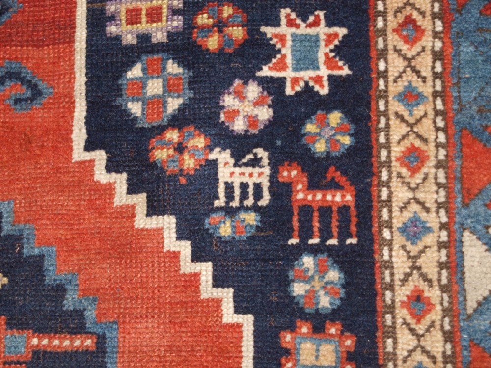 An antique hand-knotted Caucasian wool rug featuring red geometric diamond medallions on an indigo blue field with small human and animal figures. View X