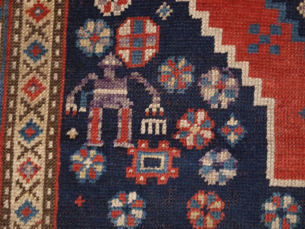 An antique hand-knotted Caucasian wool rug featuring red geometric diamond medallions on an indigo blue field with small human and animal figures. View XII