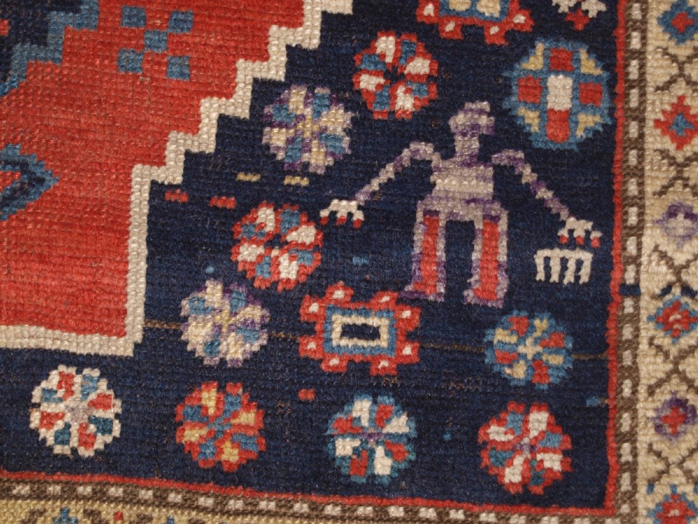 An antique hand-knotted Caucasian wool rug featuring red geometric diamond medallions on an indigo blue field with small human and animal figures. View XIII