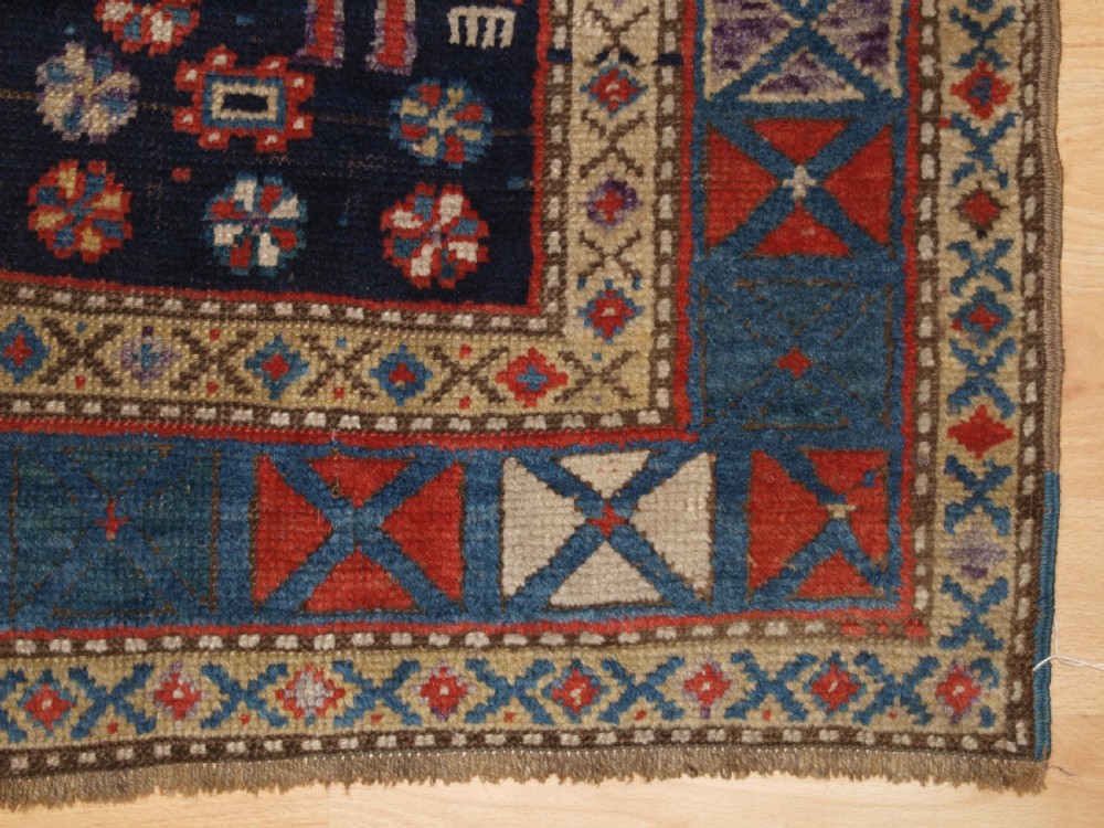 An antique hand-knotted Caucasian wool rug featuring red geometric diamond medallions on an indigo blue field with small human and animal figures. View XIV