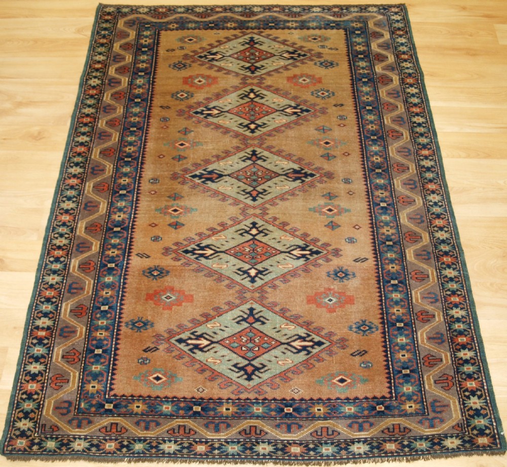 An antique Caucasian Derbent hand-knotted wool rug featuring five repeating diamond medallions in soft green and terracotta on a camel brown field with a geometric border.