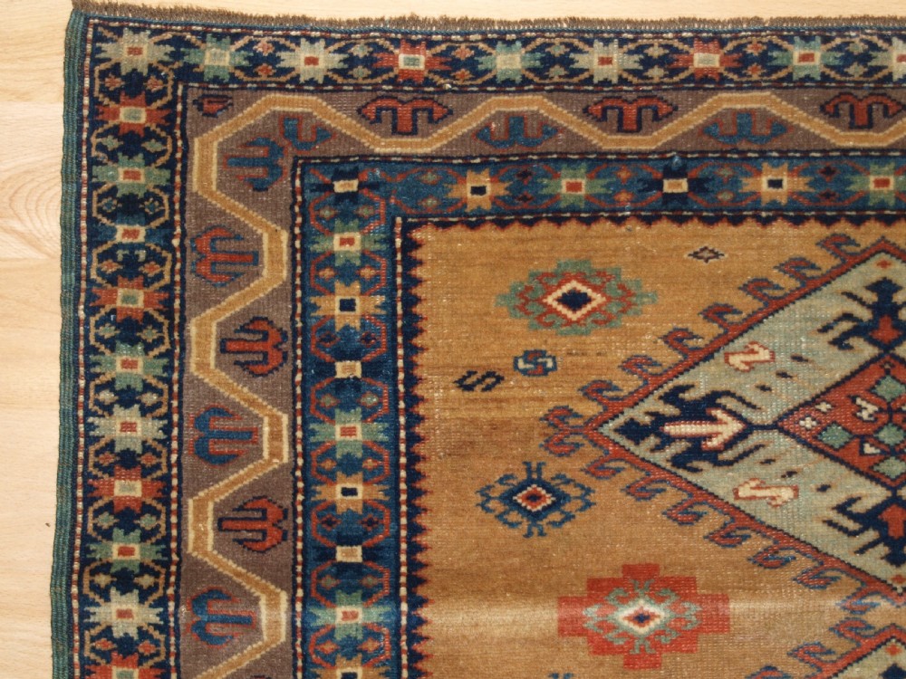 An antique Caucasian Derbent hand-knotted wool rug featuring five repeating diamond medallions in soft green and terracotta on a camel brown field with a geometric border. View III