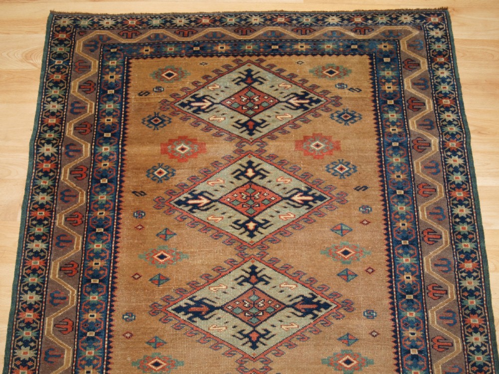 An antique Caucasian Derbent hand-knotted wool rug featuring five repeating diamond medallions in soft green and terracotta on a camel brown field with a geometric border. View IV