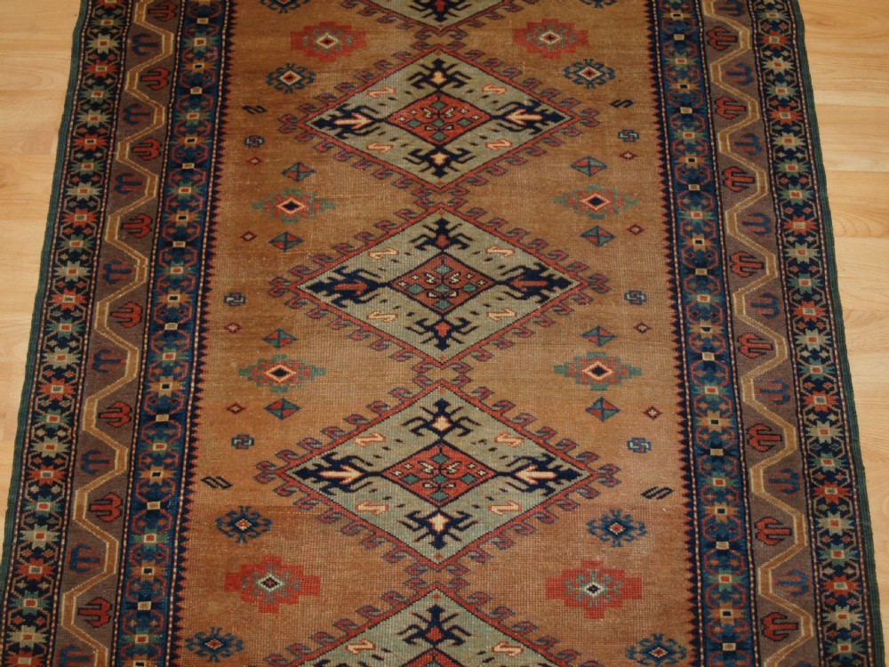 An antique Caucasian Derbent hand-knotted wool rug featuring five repeating diamond medallions in soft green and terracotta on a camel brown field with a geometric border. View V