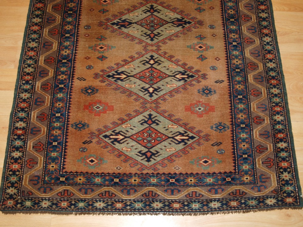 An antique Caucasian Derbent hand-knotted wool rug featuring five repeating diamond medallions in soft green and terracotta on a camel brown field with a geometric border. View VI