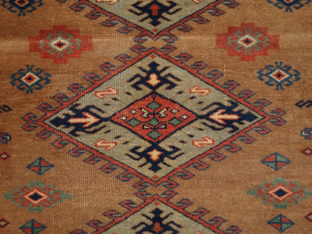 An antique Caucasian Derbent hand-knotted wool rug featuring five repeating diamond medallions in soft green and terracotta on a camel brown field with a geometric border. View VII