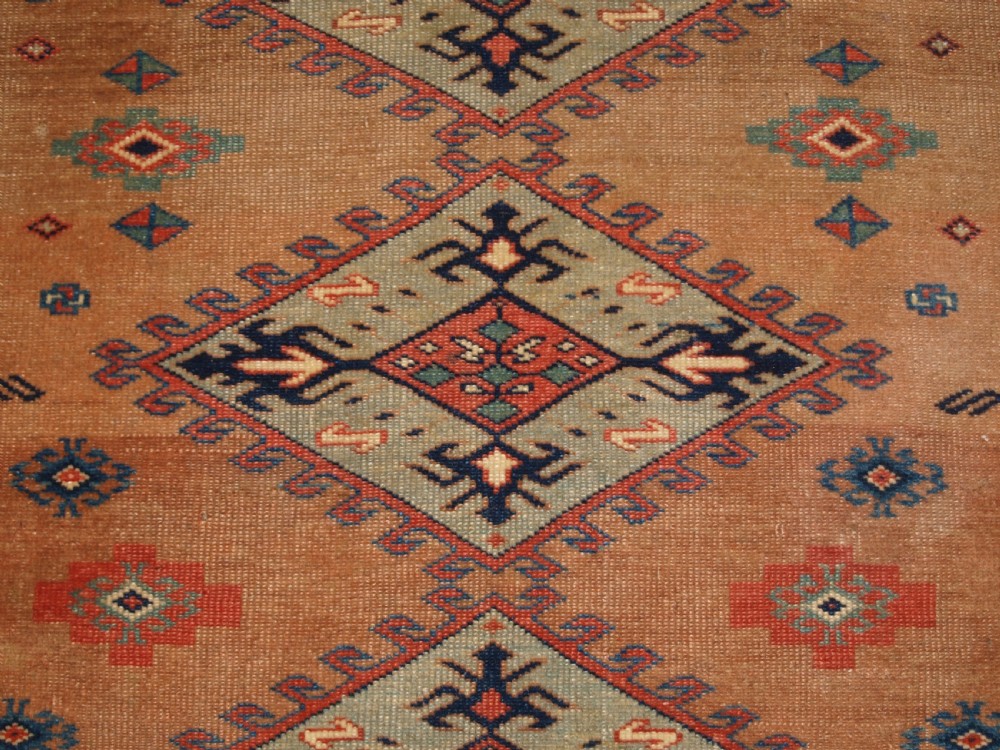 An antique Caucasian Derbent hand-knotted wool rug featuring five repeating diamond medallions in soft green and terracotta on a camel brown field with a geometric border. View IX
