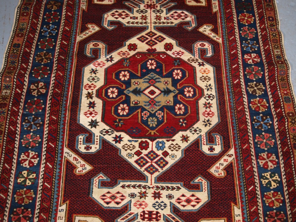 An antique Caucasian Shirvan wool rug featuring a central ivory and deep red Gymyl medallion surrounded by a dark blue rosette border. View IV