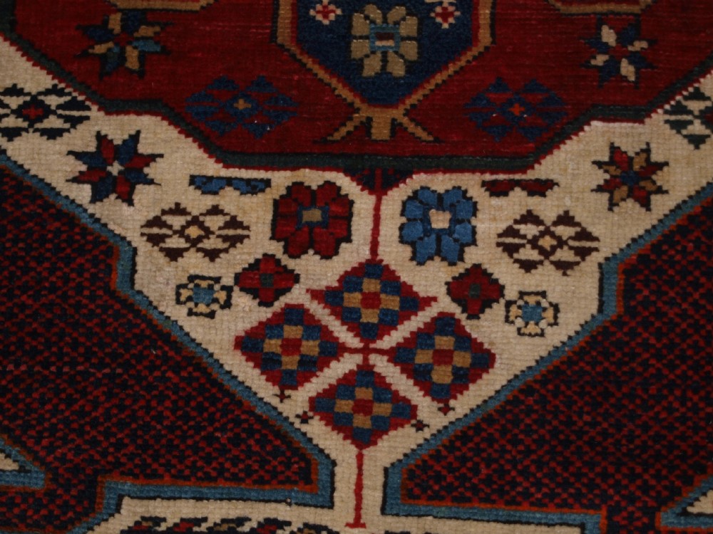 An antique Caucasian Shirvan wool rug featuring a central ivory and deep red Gymyl medallion surrounded by a dark blue rosette border. View XII