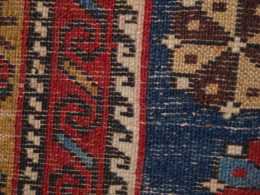 An antique Caucasian Shirvan wool rug featuring a central ivory and deep red Gymyl medallion surrounded by a dark blue rosette border. View XV