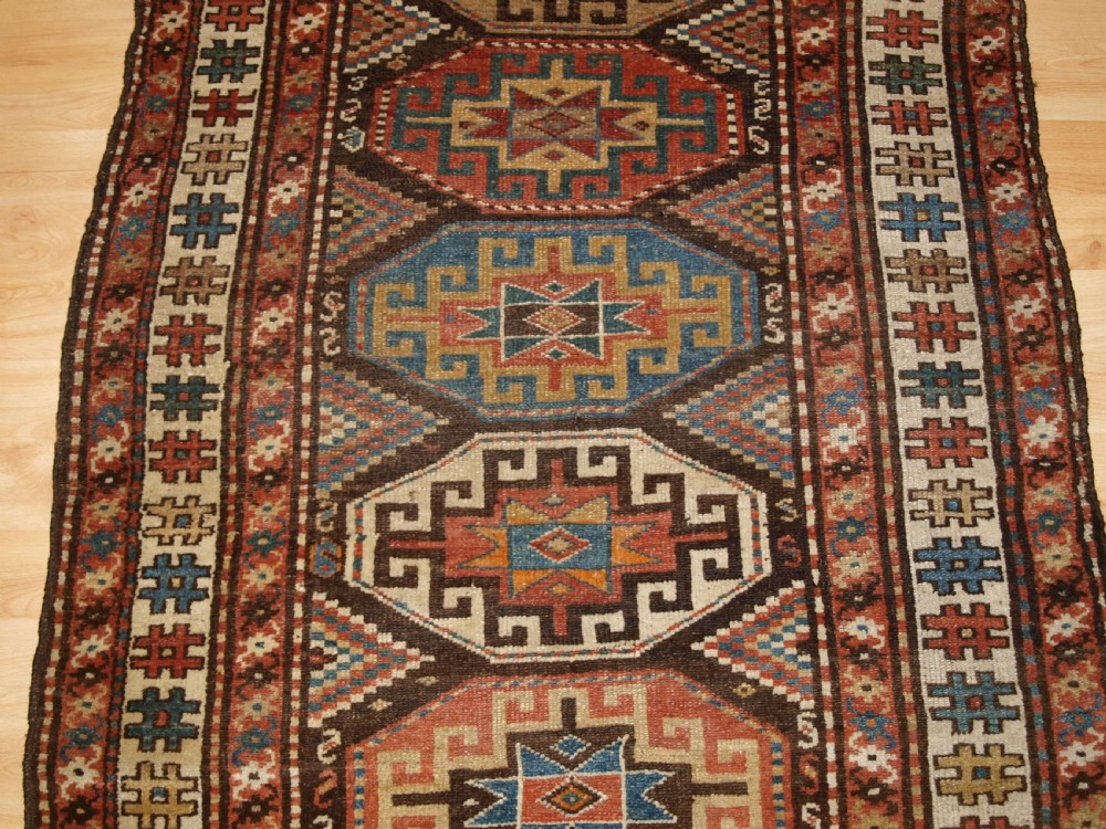 An antique hand-knotted Kurdish runner rug featuring a vertical sequence of octagonal Memling gul motifs in terracotta, blue, and cream shades. View IV