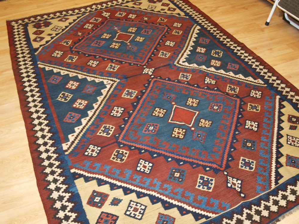 An antique Persian Qashqai flatweave wool kilim rug featuring two central blue and red diamond medallions framed by geometric tribal borders. View II