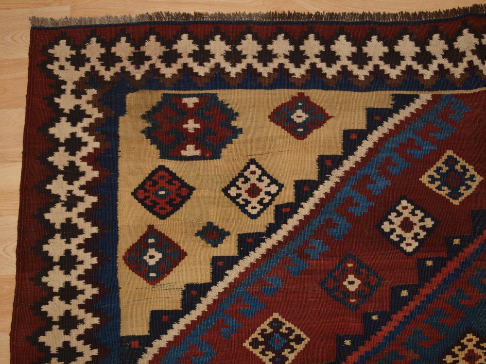 An antique Persian Qashqai flatweave wool kilim rug featuring two central blue and red diamond medallions framed by geometric tribal borders. View III