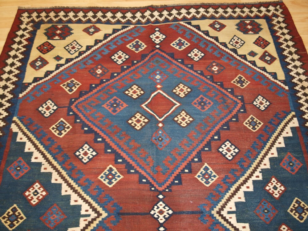 An antique Persian Qashqai flatweave wool kilim rug featuring two central blue and red diamond medallions framed by geometric tribal borders. View IV