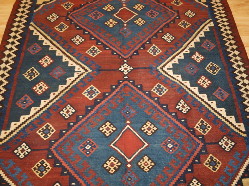 An antique Persian Qashqai flatweave wool kilim rug featuring two central blue and red diamond medallions framed by geometric tribal borders. View V