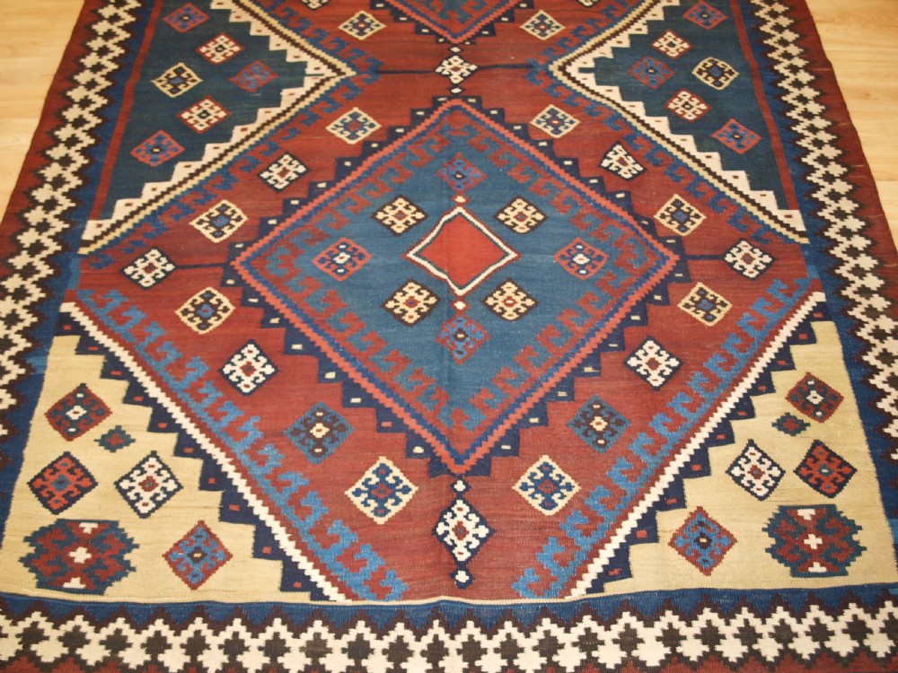 An antique Persian Qashqai flatweave wool kilim rug featuring two central blue and red diamond medallions framed by geometric tribal borders. View VI