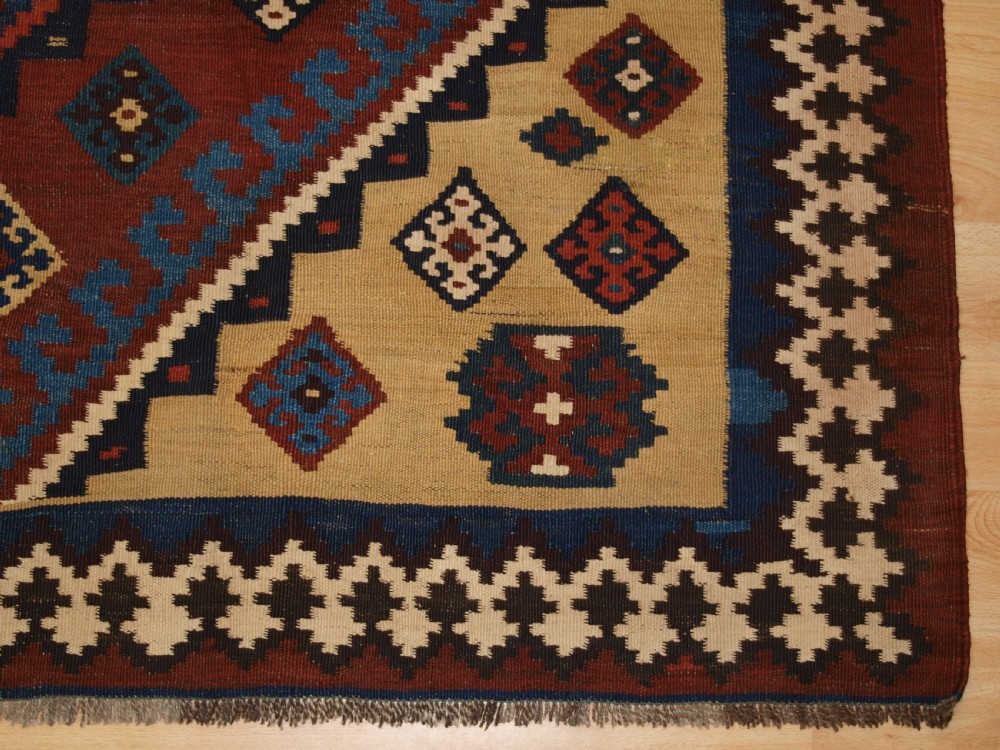 An antique Persian Qashqai flatweave wool kilim rug featuring two central blue and red diamond medallions framed by geometric tribal borders. View X