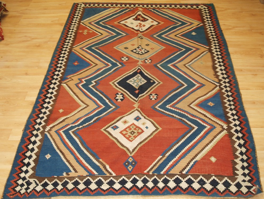 An antique flatweave Qashqai wool kilim rug featuring bold geometric diamond and chevron eye-dazzler patterns in terracotta red, indigo blue, cream, and brown.