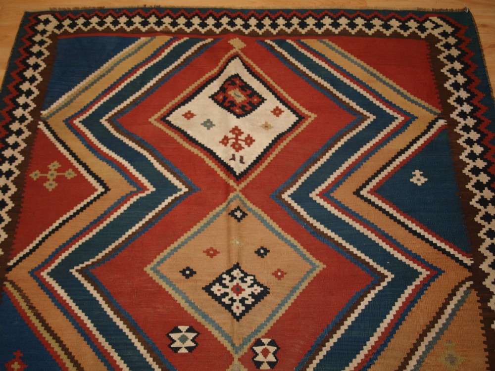 An antique flatweave Qashqai wool kilim rug featuring bold geometric diamond and chevron eye-dazzler patterns in terracotta red, indigo blue, cream, and brown. View III