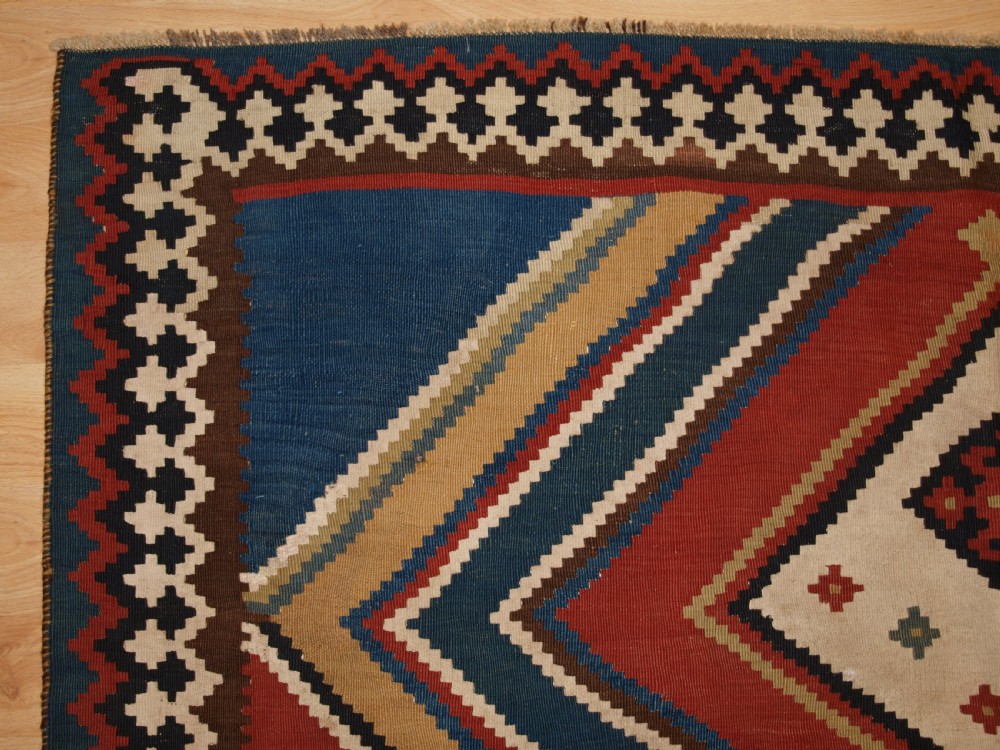 An antique flatweave Qashqai wool kilim rug featuring bold geometric diamond and chevron eye-dazzler patterns in terracotta red, indigo blue, cream, and brown. View IV
