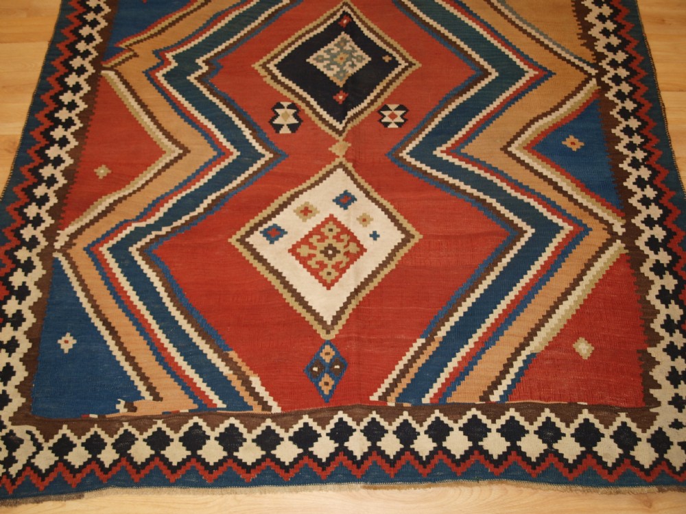 An antique flatweave Qashqai wool kilim rug featuring bold geometric diamond and chevron eye-dazzler patterns in terracotta red, indigo blue, cream, and brown. View V