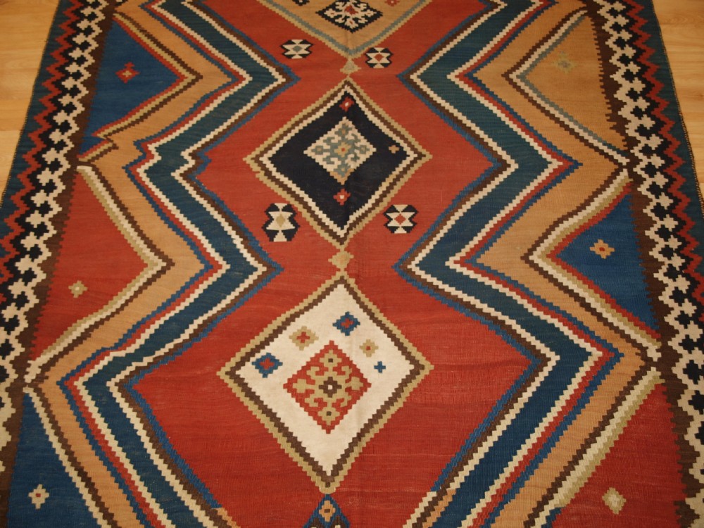 An antique flatweave Qashqai wool kilim rug featuring bold geometric diamond and chevron eye-dazzler patterns in terracotta red, indigo blue, cream, and brown. View VI
