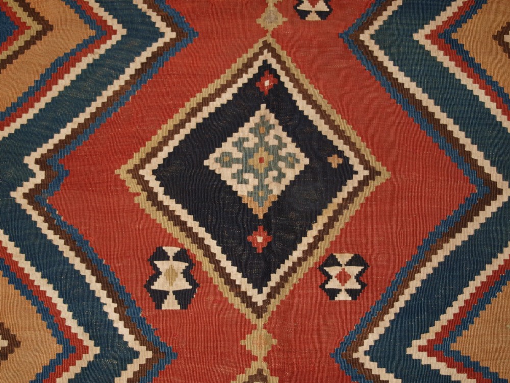 An antique flatweave Qashqai wool kilim rug featuring bold geometric diamond and chevron eye-dazzler patterns in terracotta red, indigo blue, cream, and brown. View X