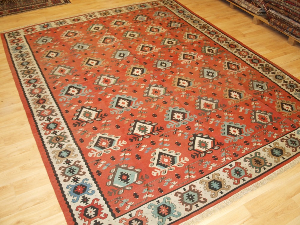 An antique flatwoven wool Sharkoy kilim rug featuring a terracotta red field with repeating geometric diamond motifs in pastel shades and a wide geometric border.