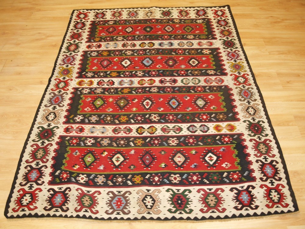 A rectangular handwoven antique wool kilim rug featuring horizontal red and dark brown banded panels decorated with geometric diamond motifs, framed by a broad cream border.