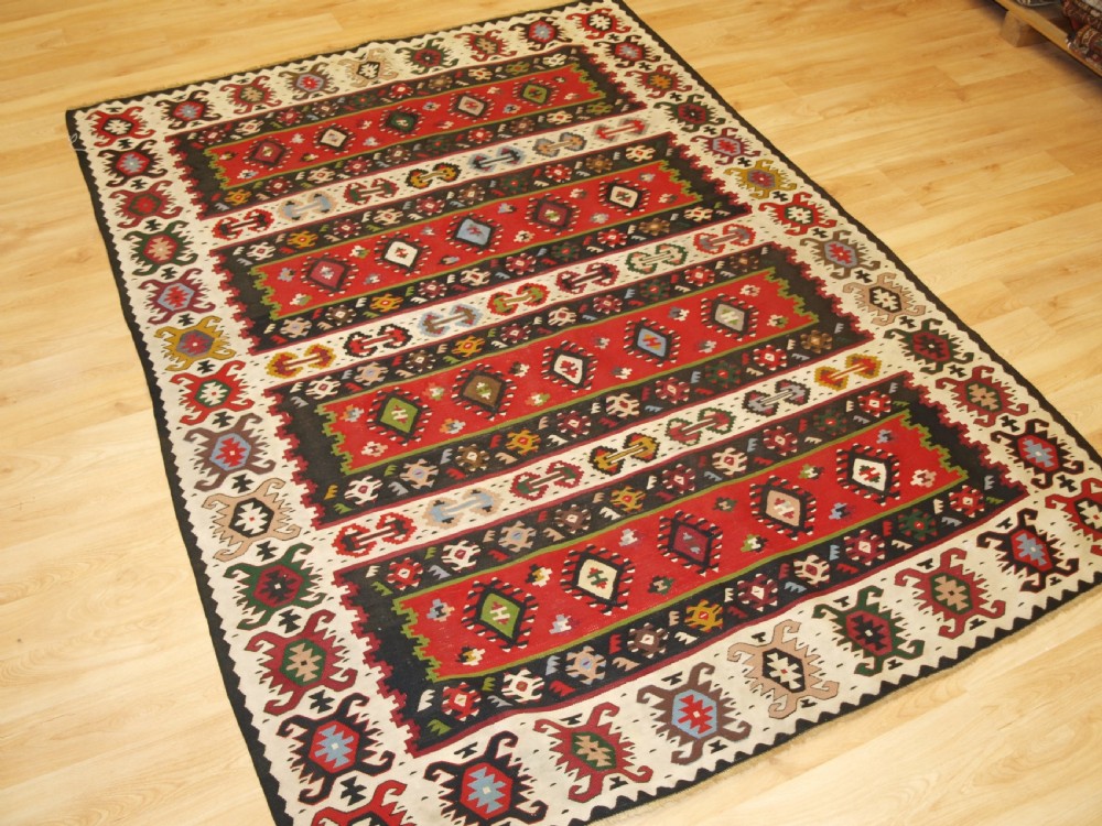 A rectangular handwoven antique wool kilim rug featuring horizontal red and dark brown banded panels decorated with geometric diamond motifs, framed by a broad cream border. View II