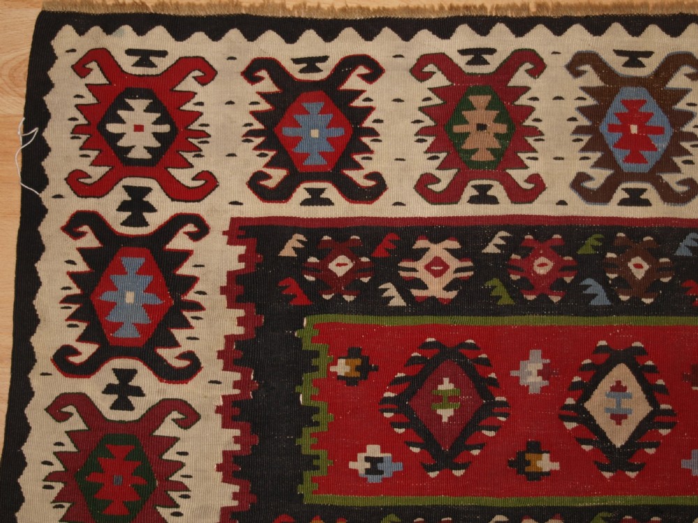 A rectangular handwoven antique wool kilim rug featuring horizontal red and dark brown banded panels decorated with geometric diamond motifs, framed by a broad cream border. View III