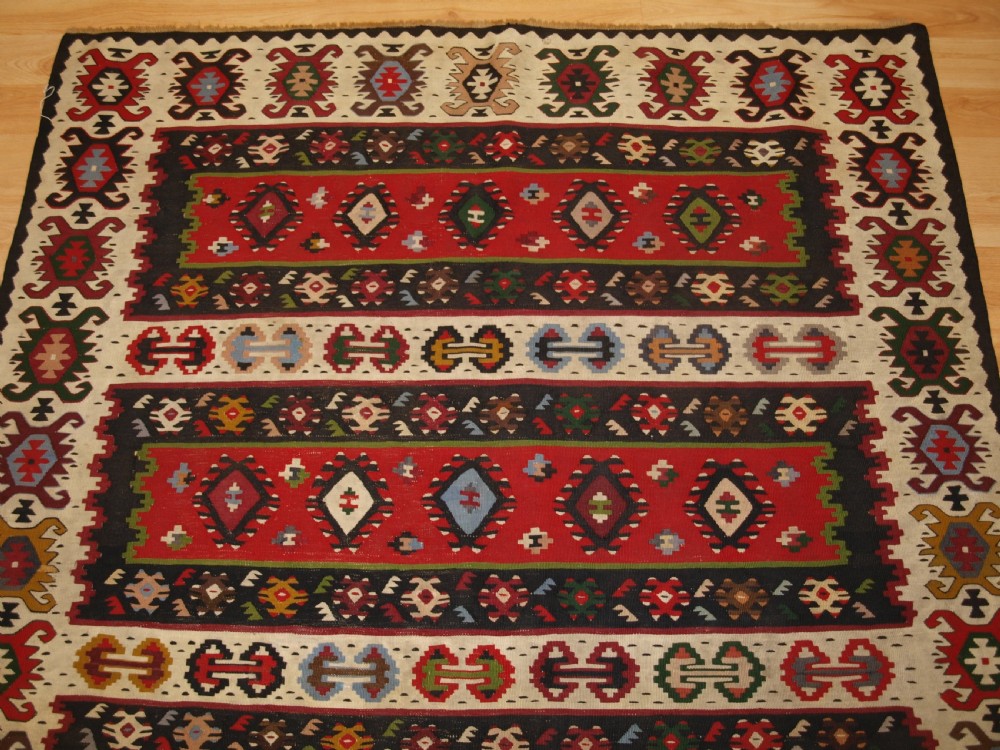 A rectangular handwoven antique wool kilim rug featuring horizontal red and dark brown banded panels decorated with geometric diamond motifs, framed by a broad cream border. View IV