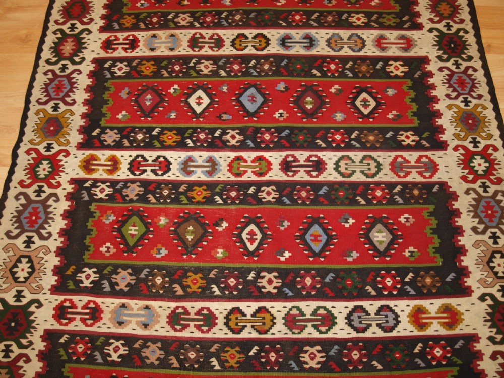 A rectangular handwoven antique wool kilim rug featuring horizontal red and dark brown banded panels decorated with geometric diamond motifs, framed by a broad cream border. View V